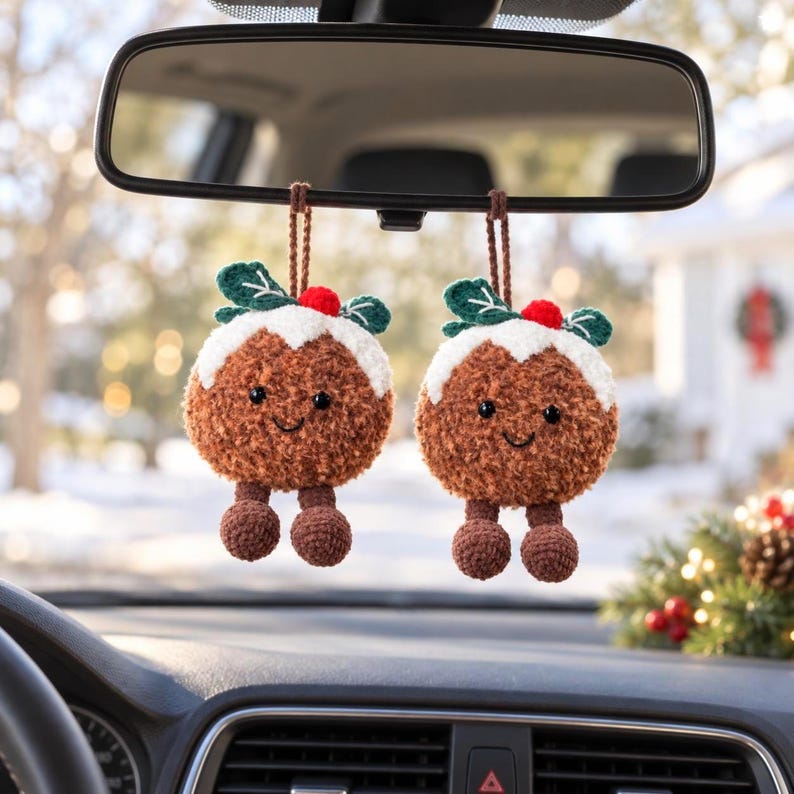 May include: Two brown crocheted Christmas pudding ornaments hanging from a rearview mirror. Each plush pudding features a white frosting top, green leaves, a red berry, black bead eyes, a stitched smile, and two dangling brown feet.