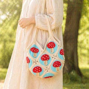 Pode incluir: Uma bolsa tote de crochê feita à mão construída com motivos quadrados apresentando um centro circular azul claro com um design de cogumelo vermelho e branco, bordado com fio cor de creme, com duas alças de ombro cor de creme.