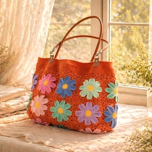 May include: An orange crochet tote bag with brown leather handles. The bag is decorated with colourful crochet flowers in shades of pink, blue, green, and purple. The bag is on a table near a window.