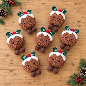 May include: Six crocheted Christmas pudding ornaments with brown textured bodies, white icing tops, green leaves, and small red berries. Each pudding features a smiling face with black eyes and two small brown dangling feet.