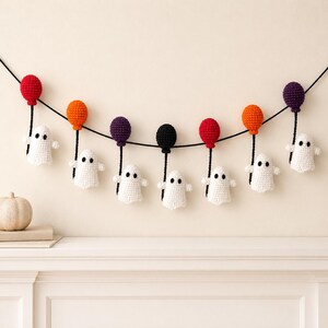 May include: A decorative garland featuring seven crocheted white ghosts, each holding a string attached to a crocheted balloon. The balloons are coloured red, orange, purple, and black, all suspended from a single black cord.