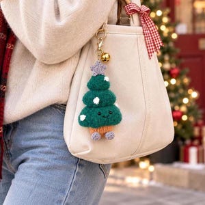 May include: A cream-colored canvas tote bag featuring a gold-toned metal keychain with a crocheted green Christmas tree charm. The tree has a small purple star on top, white embroidered snow accents, a smiling face, a brown trunk, and purple feet.