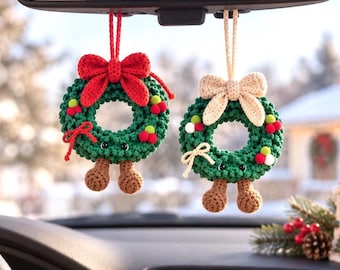 May include: Two crocheted Christmas wreath ornaments hanging from a rearview mirror. Each green wreath features small black eyes, brown feet, a crocheted bow, and tiny red and white berry accents. One wreath has a red bow, the other a beige bow.