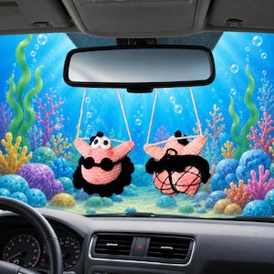 May include: Two crocheted pink starfish ornaments hanging from a rearview mirror. Each figure features a black bikini top and a black skirt, with one figure showing a black fishnet overlay on the skirt. The figures have white eyes and black pupils.