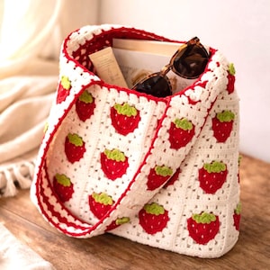 May include: A handmade crochet tote bag featuring a repeating pattern of red strawberries with green leaves on a white background. The bag has a red trim and handle, and contains sunglasses and other items. The bag is sitting on a wooden surface.