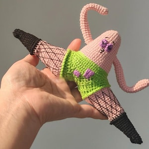 May include: A crocheted plush figure of a pink starfish character wearing bright green shorts with purple flower accents and black fishnet stockings with solid black cuffs. The figure is posed in a split with arms raised and a stitched facial expression.