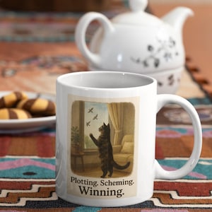 May include: White ceramic mug with a wraparound illustration of a black cat reaching for birds outside a window. The mug has the text "Plotting. Scheming. Winning." A white teapot and cookies are in the background.