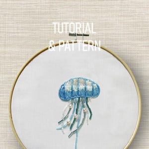 Pode incluir: Um bastidor de bordado feito à mão com um desenho de água-viva em contas nas cores azul, branco e verde. A água-viva está centrada em tecido branco, emoldurada por um aro de madeira. O texto "TUTORIAL & PATTERN" está no topo.