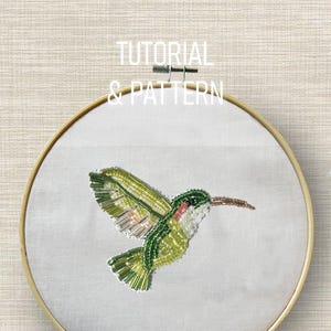 May include: A hand-stitched hummingbird embroidery on white fabric, secured in a wooden hoop. The bird features green, pink, and silver beads, with a red throat and brown beak. The text "TUTORIAL & PATTERN" is above the hoop.