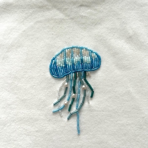 May include: Embroidered jellyfish patch in shades of blue, white, and pearl beads. The jellyfish design is on a white fabric, showcasing the detailed beadwork and aquatic theme. The patch is likely for clothing or accessories.