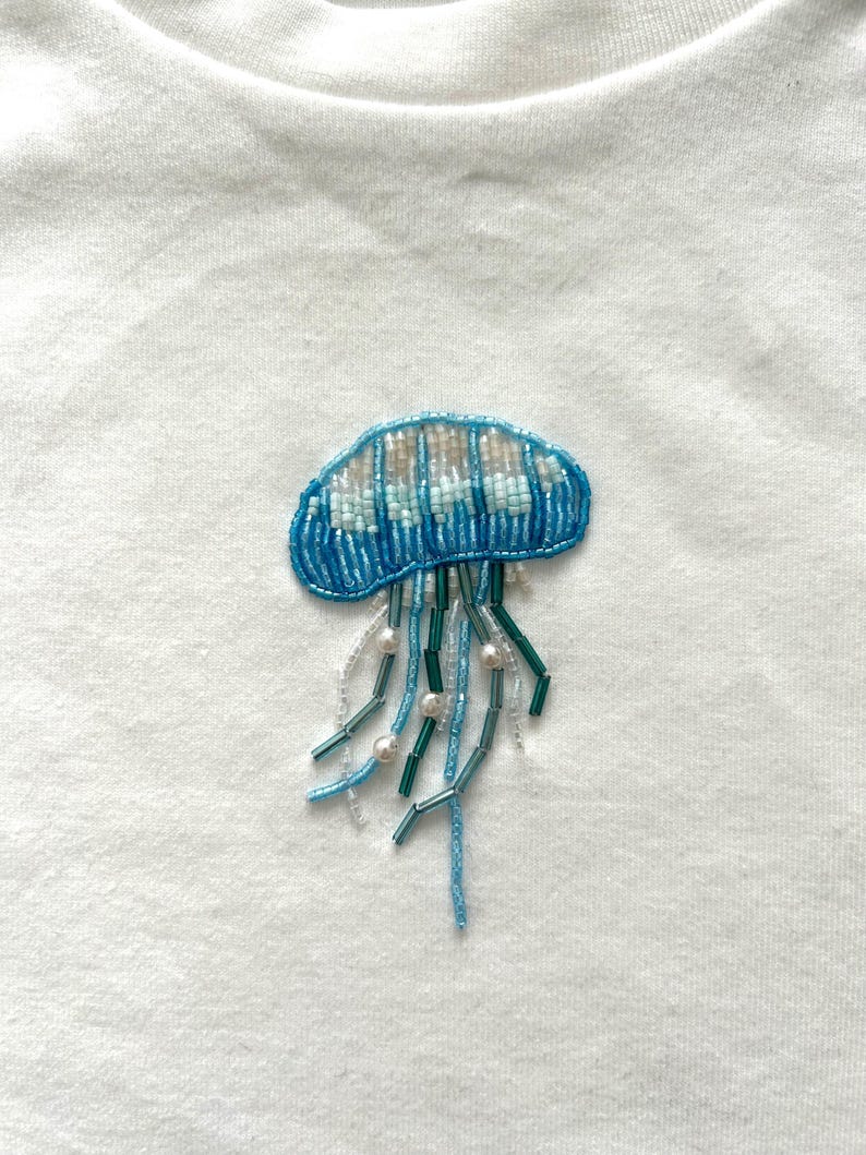 May include: Embroidered jellyfish patch in shades of blue, white, and pearl beads. The jellyfish design is on a white fabric, showcasing the detailed beadwork and aquatic theme. The patch is likely for clothing or accessories.