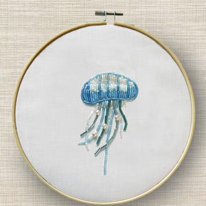 May include: A beaded jellyfish embroidery on white fabric, set in a wooden hoop. The jellyfish is blue and white, with long, flowing tentacles in shades of blue, green, and white, accented with pearl beads. The background is a light beige.