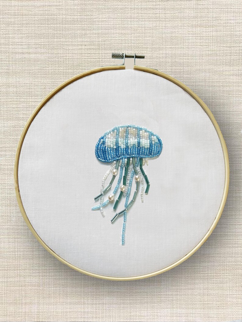 May include: A beaded jellyfish embroidery on white fabric, set in a wooden hoop. The jellyfish is blue and white, with long, flowing tentacles in shades of blue, green, and white, accented with pearl beads. The background is a light beige.