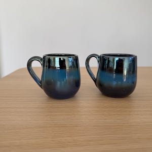 Może przedstawiać: Dwa małe, ciemnoniebieskie ceramiczne kubki o zaokrąglonych korpusach i zakrzywionych uchwytach. Kubki mają błyszczące, opalizujące wykończenie, które odbija światło, tworząc gradient niebieskich i czarnych odcieni. Stoją na jasnobrązowej drewnianej powierzchni.