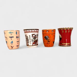 Op de afbeelding: Vier decoratieve shotglaasjes met Egyptische thema's. De glazen hebben verschillende kleuren, waaronder perzik, wit, oranje en rood. Elk glas heeft unieke illustraties, waaronder hiërogliefen, figuren en het woord "EGYPT".