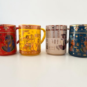 Könnte beinhalten: Vier Keramikbecher mit ägyptischen Motiven. Die Becher sind rot, gelb, beige und dunkelblau und zeigen Bilder von Pharaonen, Hieroglyphen und anderen altägyptischen Motiven. Goldakzente zieren die Ränder und Henkel.