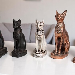 Op de afbeelding: Vijf Egyptische kattenbeelden in verschillende kleuren, waaronder zwart, wit en brons. De beelden staan in een zittende positie, met gedetailleerde kragen en sokkels. De beelden worden tentoongesteld op een witte tafel.
