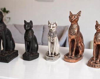 May include: Five Egyptian cat statues in varying colors, including black, white, and bronze. The statues are in a seated position, with detailed collars and bases. The statues are displayed on a white table.