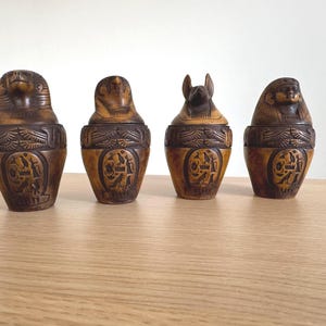 Può includere: Quattro urne decorative in legno in stile egizio con dettagli intagliati. Ogni urna ha una testa di animale o umana diversa sul coperchio. Le urne sono marroni con geroglifici intagliati e fasce decorative.