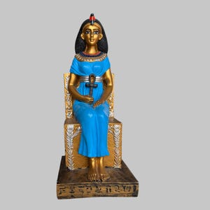 Può includere: Statua dorata di una regina egizia vestita con una tunica blu e una corona d'oro, che tiene un ankh. La statua è seduta su un trono con dettagli dorati e argentati. La base della statua è decorata con geroglifici.