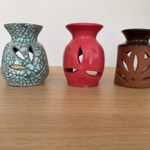 Op de afbeelding: Drie keramische vazen in verschillende kleuren. De eerste is groen met een craquelé patroon, de tweede is rood en de derde is bruin met bladvormige uitsparingen. Ze hebben allemaal openingen.