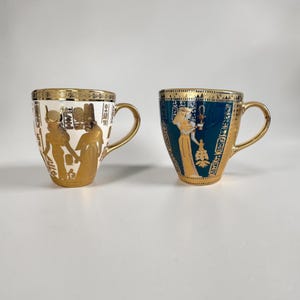 Pode incluir: Duas canecas de cerâmica com detalhes dourados. Uma é branca com hieróglifos e figuras douradas. A outra é turquesa com hieróglifos e uma figura dourada. Ambas as canecas têm bordas e alças douradas, com tema do antigo Egito.