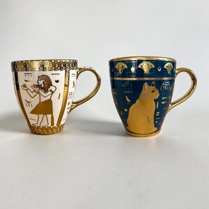 Könnte beinhalten: Zwei Keramikbecher mit goldenen Akzenten und Griffen. Ein Becher ist weiß mit goldenen Hieroglyphen und einer Figur. Der andere ist teal mit einer goldenen Katzenillustration und Hieroglyphen. Beide Becher haben goldene Ränder.