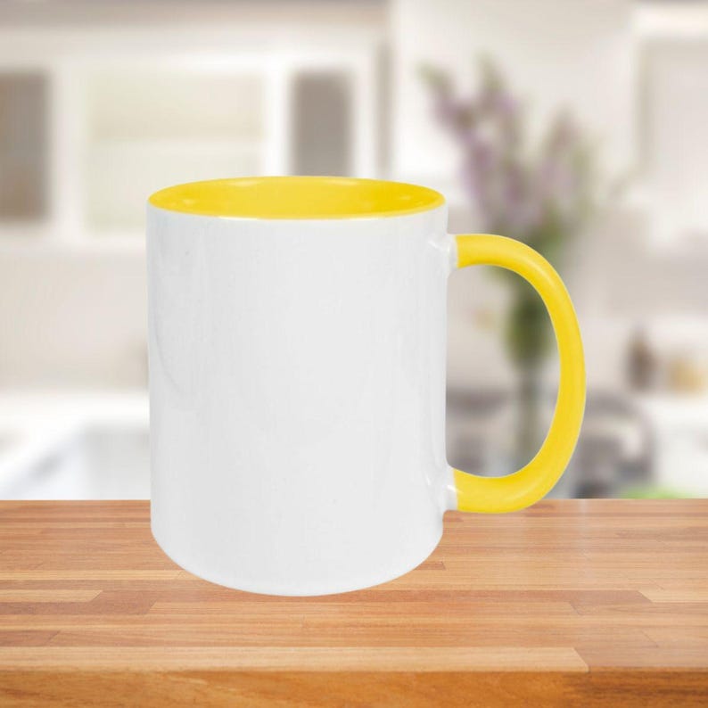 May include: A white ceramic mug with a yellow interior and handle. The mug is sitting on a wooden surface. The background is blurred, suggesting a kitchen setting.