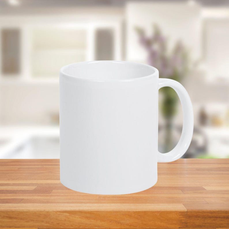 May include: A white ceramic mug with a curved handle. The mug is cylindrical and sits on a wooden surface. The background is blurred, suggesting a kitchen or dining setting. The mug is empty.