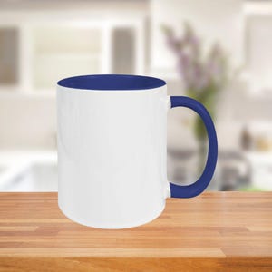 May include: A white ceramic mug with a blue interior, handle, and rim. The mug is cylindrical and sits on a wooden surface. The background is a blurred kitchen scene.