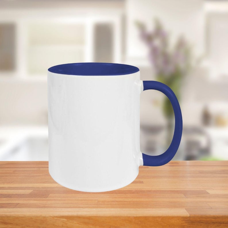 May include: A white ceramic mug with a blue interior, handle, and rim. The mug is cylindrical and sits on a wooden surface. The background is a blurred kitchen scene.