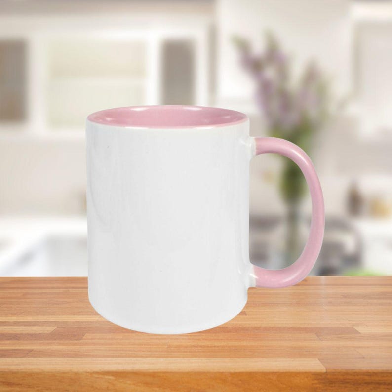 May include: A white ceramic mug with a pink interior and handle. The mug is set on a wooden surface, with a blurred background of a kitchen setting.