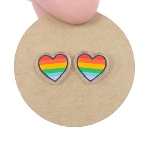 May include: Two heart-shaped earrings with a rainbow design, set on a round, tan cardboard backing. Each earring features a clear border and a vibrant spectrum of colors, including red, orange, yellow, green, blue, and purple.