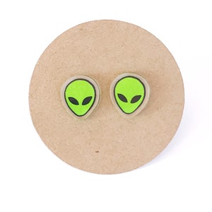 May include: A pair of green alien face stud earrings. The earrings are made of clear plastic with a green alien face printed on them.
