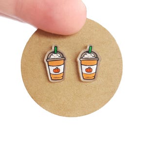 May include: A pair of clear acrylic earrings featuring a cartoon design of a pumpkin spice latte. The earrings have a white whipped cream topping, an orange cup with a pumpkin, and a green straw. The earrings are on a brown circular surface.