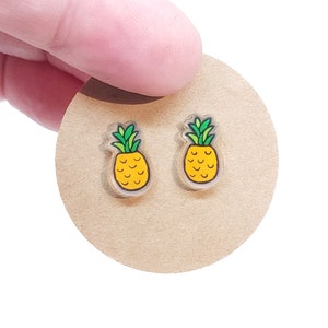 May include: A pair of clear acrylic pineapple stud earrings. Each earring features a yellow pineapple with green leaves and a yellow border. The earrings are displayed on a tan circular backing.