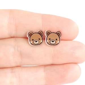 May include: A pair of small, clear acrylic bear-shaped earrings. Each earring features a brown bear face with black outlines, small black eyes, and a light brown nose. The earrings are held in a person's hand.