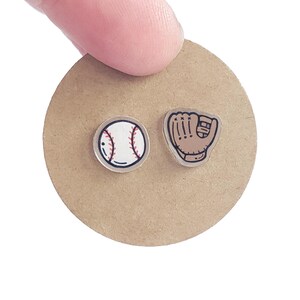 May include: Clear acrylic baseball earrings. One earring is a baseball with red stitching, and the other is a baseball glove. The earrings are on a round, tan cardboard backing.