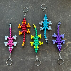 Op de afbeelding: Vijf kleurrijke kralen-hagedis sleutelhangers. De hagedissen zijn roze, rood, geel, groen en paars. Elke sleutelhanger heeft een zilveren metalen ring.