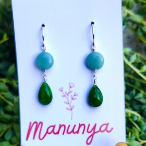 May include: A pair of dangle earrings with a teal round bead and a green teardrop-shaped bead. The earrings are displayed on a white card with the name "Manunya" in pink script. The background is a lush green plant.