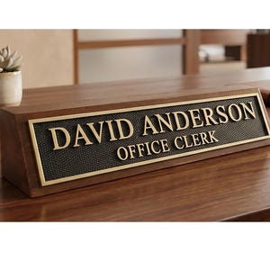 Könnte beinhalten: Ein dreieckiges Schreibtisch-Namensschild aus Holz mit dunkelbraunem Finish und einer rechteckigen Metallplatte. Die Platte hat einen strukturierten schwarzen Hintergrund mit goldfarbener Reliefschrift: DAVID ANDERSON OFFICE CLERK.