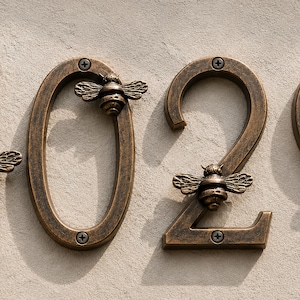 Może przedstawiać: Zestaw czterech numerów domu wyświetlających 1029, wykonanych z ciemnego metalu w kolorze brązu z wykończeniem typu weathered. Każda cyfra posiada małe, szczegółowe zdobienia w kształcie pszczół, przymocowane widocznymi śrubami w punktach montażowych.