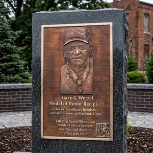 May include: A rectangular bronze plaque mounted on a dark gray granite stone monument. The plaque features a relief portrait of Gary G. Wetzel wearing a U.S. Army Veteran cap and a Medal of Honor, with engraved text and a QR code in the bottom corner.