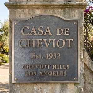 May include: A rectangular bronze-toned metal plaque with an arched top, mounted with four screws. The plaque features raised lettering that reads CASA DE CHEVIOT, Est. 1932, CHEVIOT HILLS, and LOS ANGELES, set against a dark, textured background.