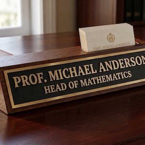 Peut inclure: Un porte-nom de bureau triangulaire en bois foncé avec une plaque métallique noire et des lettres dorées indiquant PROF. MICHAEL ANDERSON HEAD OF MATHEMATICS. La base comporte une fente contenant des cartes de visite avec un logo doré.