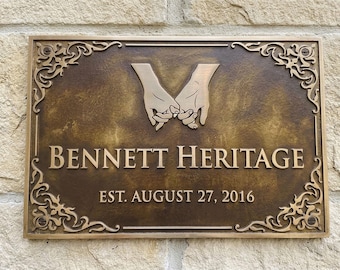 May include: A rectangular bronze-colored metal plaque featuring a raised relief of two hands interlocking pinky fingers at the top. The text reads BENNETT HERITAGE and EST. AUGUST 27, 2016. The border has decorative scrollwork in each corner.