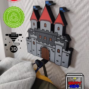 Könnte beinhalten: Ein 3D-gedruckter Wandhaken in Burgform mit roten Dachzinnen, grauen Mauern und einer braunen Holztür. Der Haken ist zum Aufhängen von Mänteln oder anderen Gegenständen gedacht. Das Bild enthält den Text "HAPPY KIDS" und "MADE IN GERMANY".