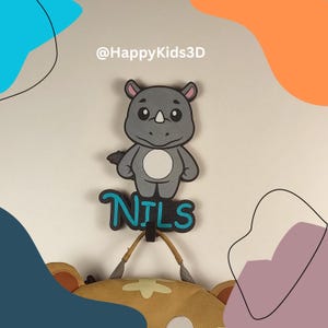 Könnte beinhalten: Ein grauer Nashorn-Wandhaken mit dem Namen "NILS" in Blau und Schwarz. Der Haken hält eine beige und braune Tasche. Der Text "@HappyKids3D" steht oben.