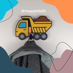 Könnte beinhalten: Ein gelb-schwarzer Wandhaken in Form eines Muldenkippers mit einer Ladung braunen Materials. Der Haken hält ein graues Kapuzenhandtuch. Der Text "@HappyKids3D" steht über dem LKW.