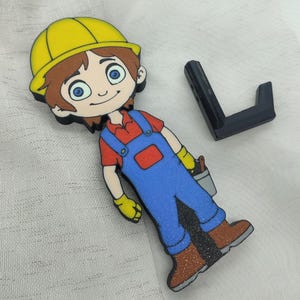 Könnte beinhalten: Eine Comicfigur eines Bauarbeiters mit gelbem Helm, rotem Hemd, blauer Latzhose und braunen Schuhen. Die Figur hält einen Pinsel und einen Eimer.